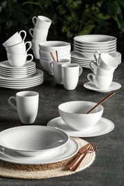 Elegantes weißes Kombiservice Beat 42-tlg. aus Porzellan, bestehend aus Tassen, Tellern und Schalen, auf einem dunklen Tisch mit grüner Pflanzenkulisse, seitliche Perspektive.