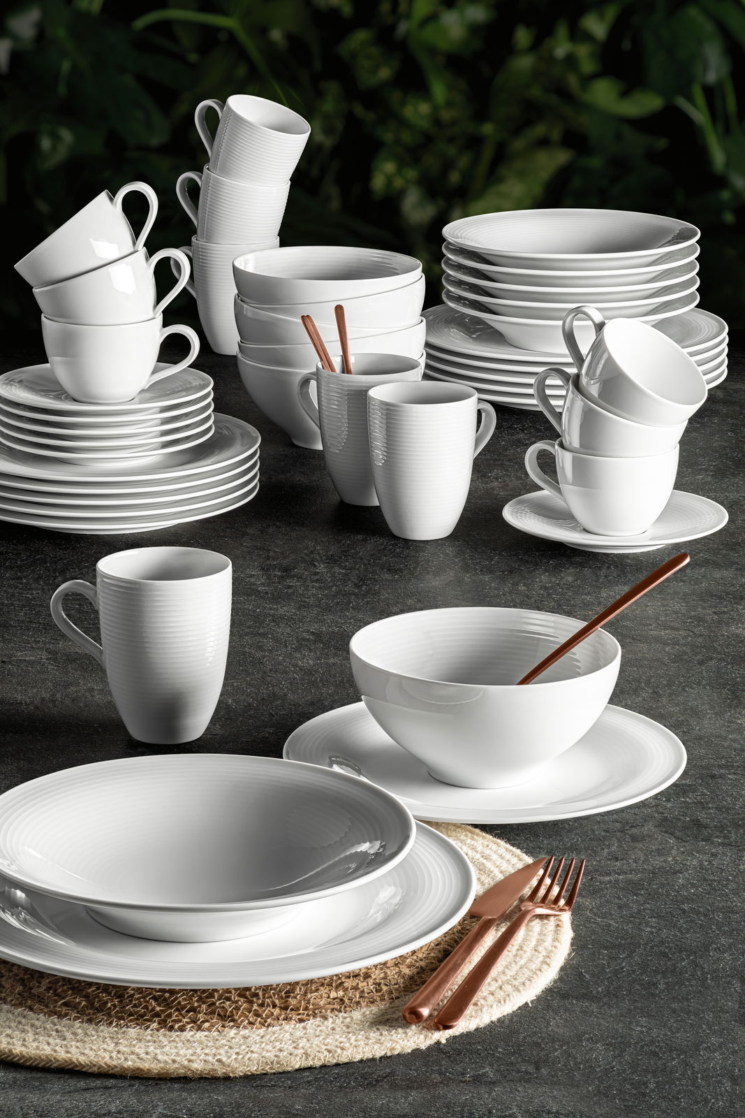 Elegantes weißes Kombiservice Beat 42-tlg. aus Porzellan, bestehend aus Tassen, Tellern und Schalen, auf einem dunklen Tisch mit grüner Pflanzenkulisse, seitliche Perspektive.
