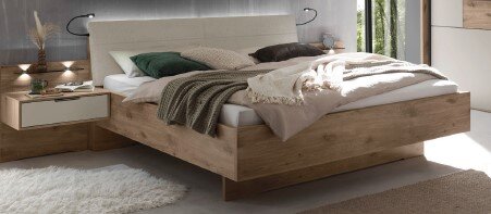 Modernes Holzbett mit gepolstertem Kopfteil in hellem Stoff, weißer Bettwäsche und Decken in Grün und Grau, im Schlafzimmer neben schwebendem Nachttisch – Perspektive: frontale 3/4-Ansicht von schräg vorne.