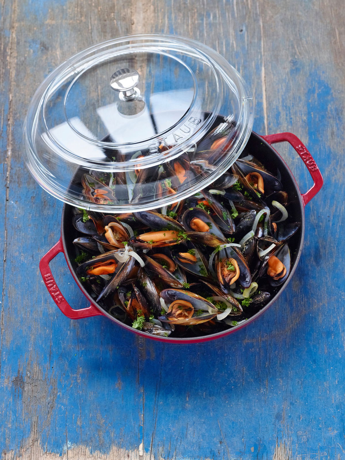 Roter Bräter mit Glasdeckel, gefüllt mit Muscheln, aus der Vogelperspektive auf einem blauen Holztisch.