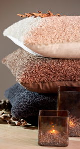 Stapel von drei Kissen in verschiedenen Farben und Texturen, von oben nach unten: beige, braun und dunkelgrau, mit dekorativen Elementen und Kerzen im Vordergrund, seitliche Perspektive.