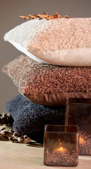 Stapel von drei Kissen in verschiedenen Farben und Texturen, von oben nach unten: beige, braun und dunkelgrau, mit dekorativen Elementen und Kerzen im Vordergrund, seitliche Perspektive.