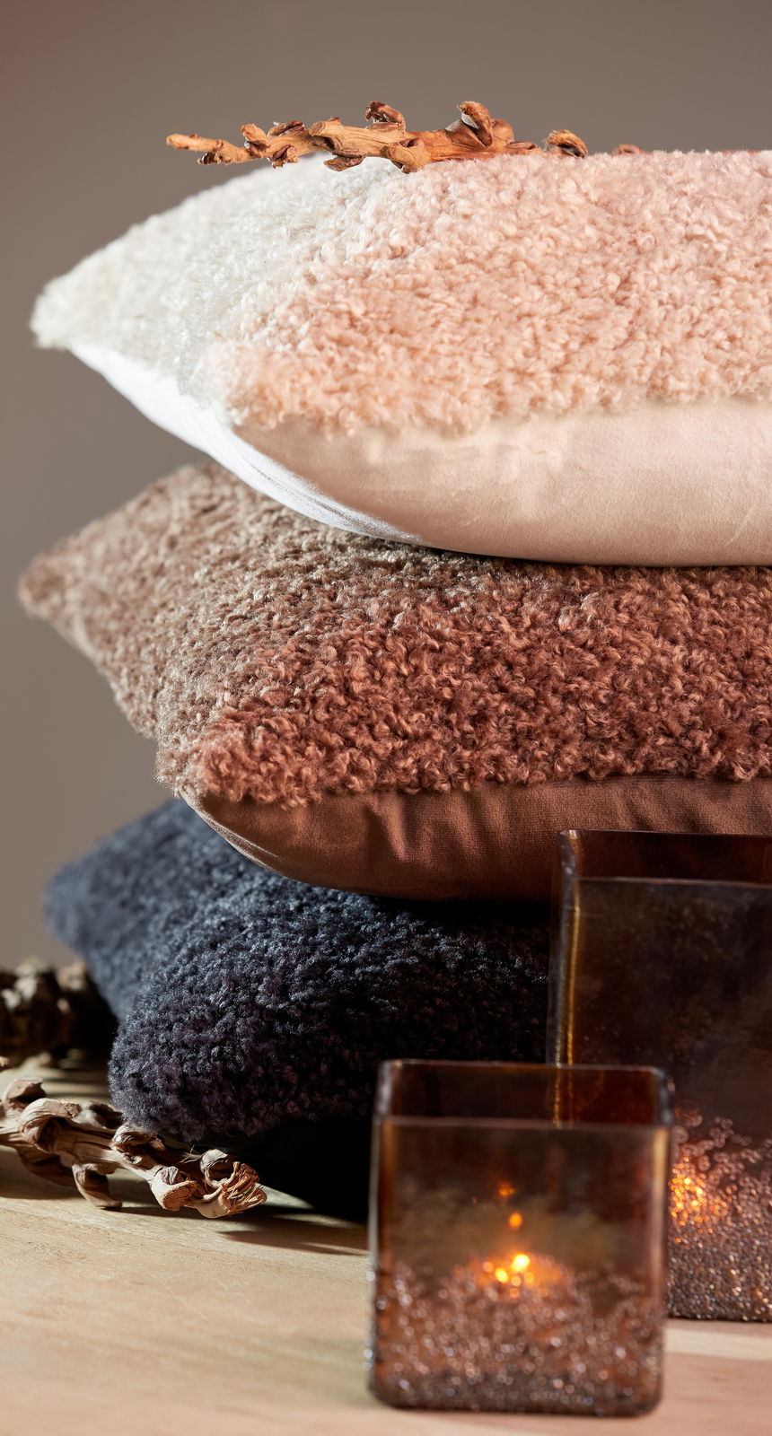 Stapel von drei Kissen in verschiedenen Farben und Texturen, von oben nach unten: beige, braun und dunkelgrau, mit dekorativen Elementen und Kerzen im Vordergrund, seitliche Perspektive.