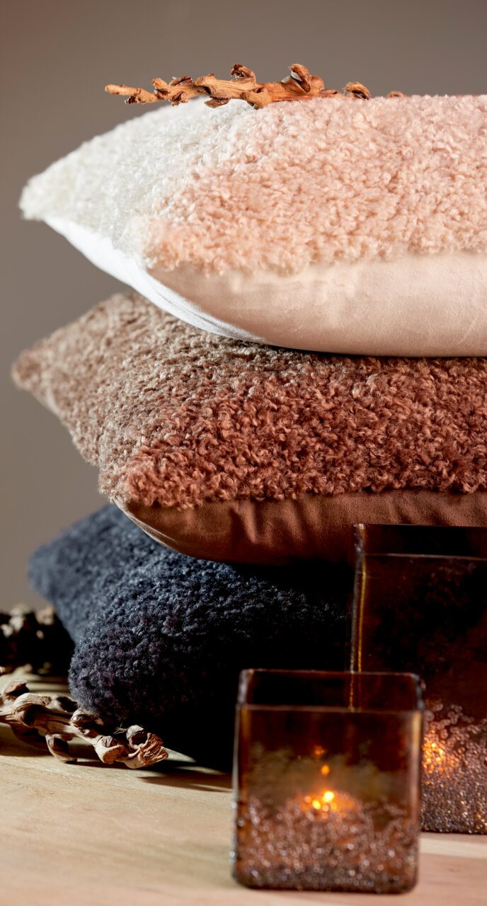 Stapel von drei Kissen in verschiedenen Farben und Texturen, von oben nach unten: beige, braun und dunkelgrau, mit dekorativen Elementen und Kerzen im Vordergrund, seitliche Perspektive.
