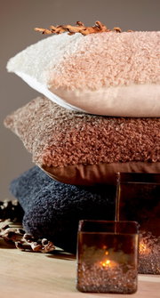 Een stapel van drie kussens in verschillende kleuren en texturen, van boven naar beneden: beige, bruin en donkergrijs, met decoratieve elementen en kaarsen op de voorgrond, gezien vanuit een zijdelingse hoek.