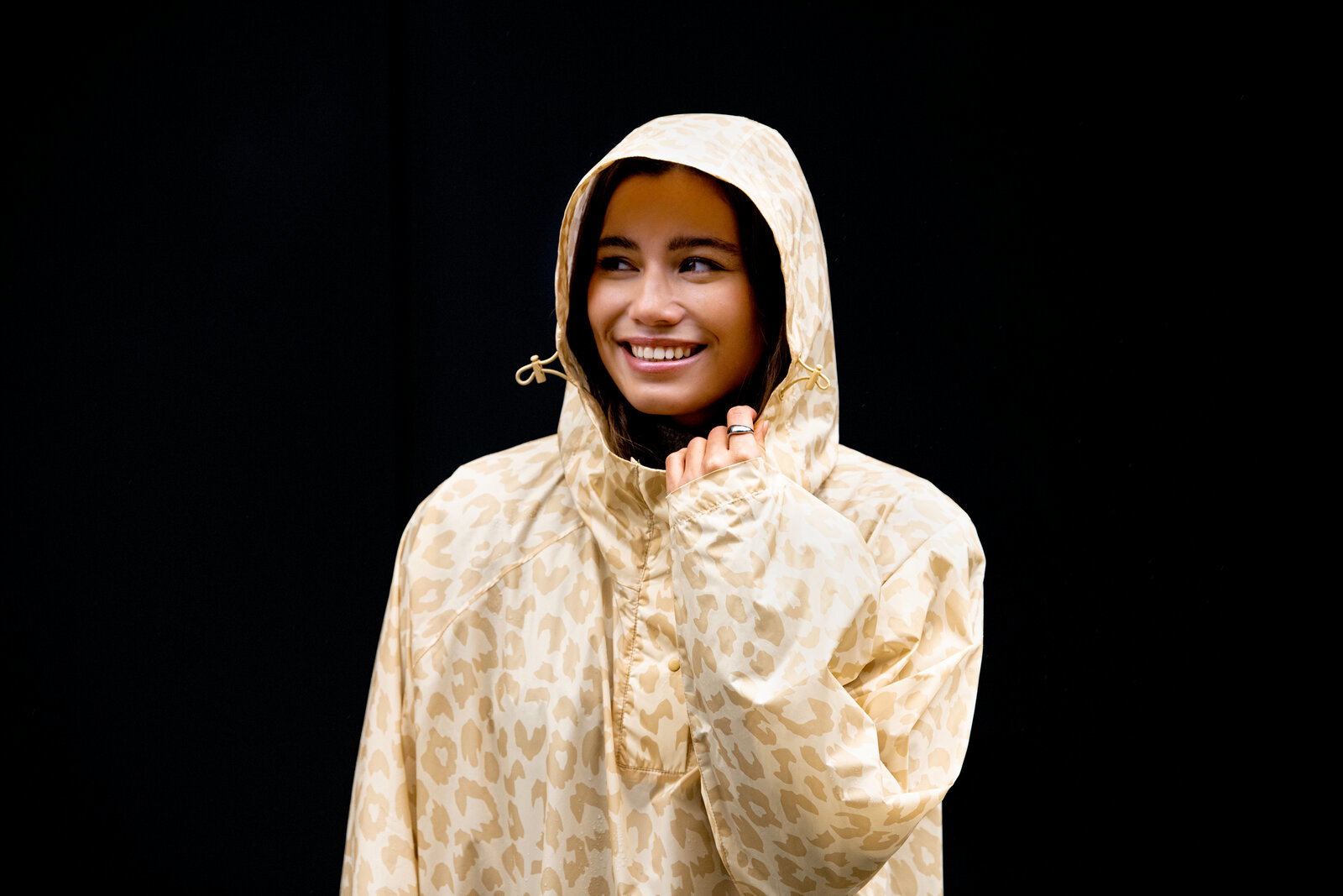 Lächelnde Frau in beigefarbener, gemusterter Regenjacke mit Kapuze und Kordelzug vor schwarzem Hintergrund, Halbporträt in leichter Seitenansicht von vorn.