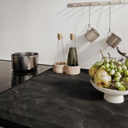 Moderne Küchenarbeitsplatte mit Induktionskochfeld, Edelstahlkochtopf, zwei Glasflaschen mit Holzuntersetzern, hängenden Tassen und einer Obstschale mit Trauben und Birnen, aus der Vogelperspektive betrachtet.