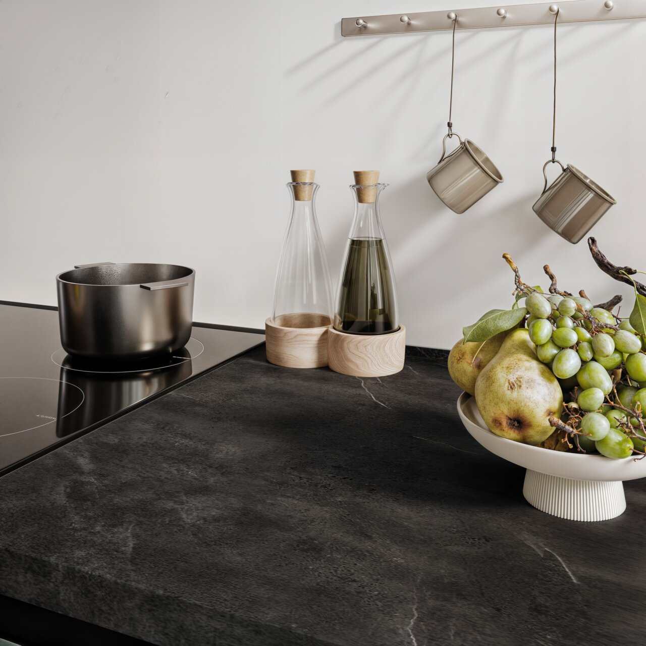 Moderne Küchenarbeitsplatte mit Induktionskochfeld, Edelstahlkochtopf, zwei Glasflaschen mit Holzuntersetzern, hängenden Tassen und einer Obstschale mit Trauben und Birnen, aus der Vogelperspektive betrachtet.