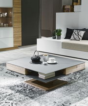 Moderner Couchtisch in quadratischer Form mit grauer Oberfläche und Holzdetails, dekoriert mit Teekanne und Tassen, seitliche Perspektive.