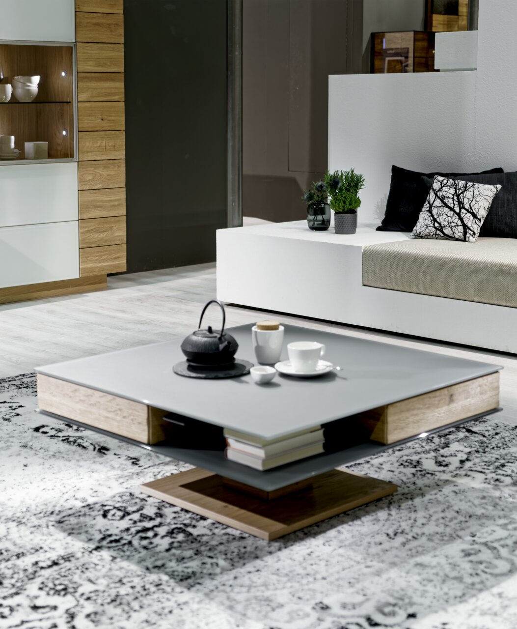 Moderner Couchtisch in quadratischer Form mit grauer Oberfläche und Holzdetails, dekoriert mit Teekanne und Tassen, seitliche Perspektive.