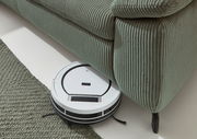 Ein Saugroboter reinigt den Boden unter einem grünen, gerippten Sofa aus der Vogelperspektive.