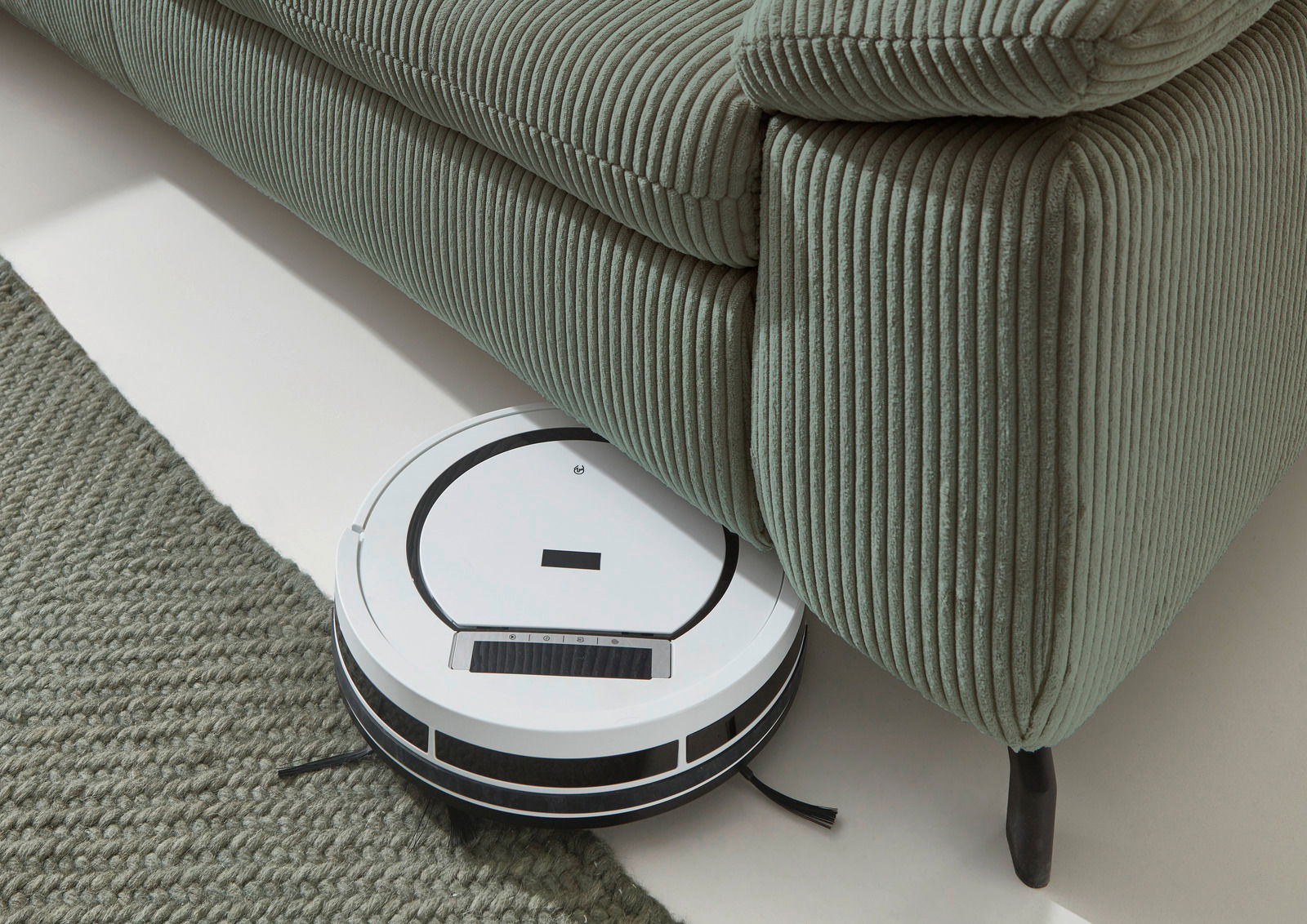Beldomo Style Ecksofa SENSE Ein Saugroboter reinigt den Boden unter einem grünen, gerippten Sofa aus der Vogelperspektive.