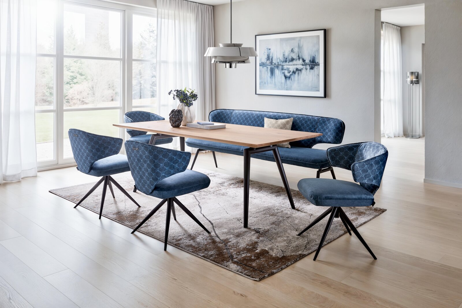 Moderne Essgruppe aus schräger Vorderansicht mit rechteckigem Holztisch, vier blauen Polsterstühlen und passender Sitzbank auf einem gemusterten Teppich