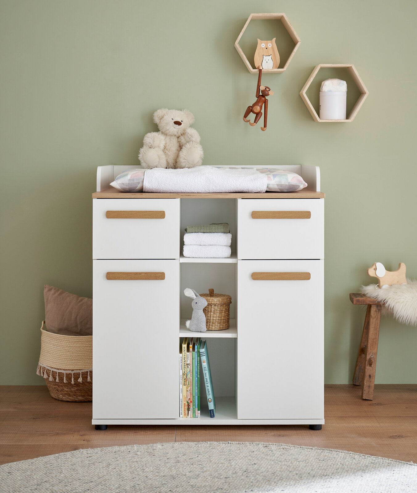 MyBaby Wickelkommode ASTEA Weiße Wickelkommode mit Holzauflage, vier Schubladen und offenen Regalfächern im Kinderzimmer, mit Wickelauflage und Teddybär obenauf; Frontalansicht auf Augenhöhe.
