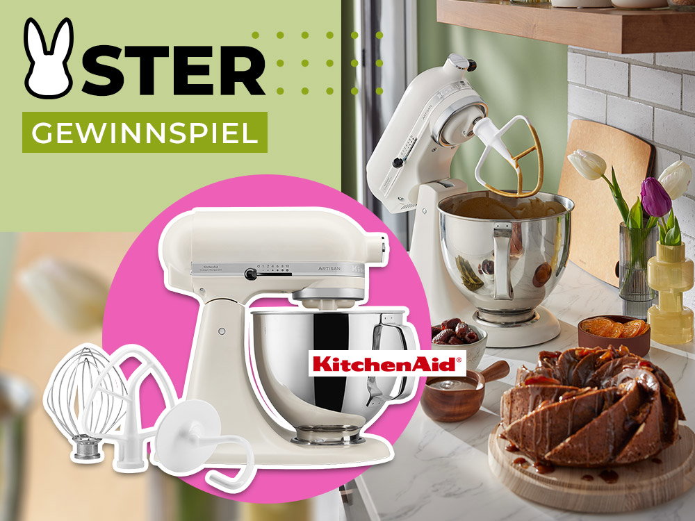 Weiße KitchenAid Küchenmaschine mit Edelstahlschüssel beim Rühren von Teig, daneben Kuchen auf Holzbrett und Tulpen in Vase