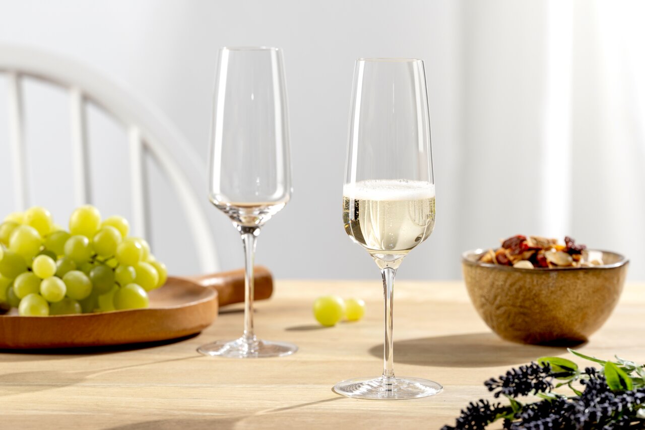 Zwei elegante Sektgläser auf einem Tisch, eines gefüllt mit Sekt, umgeben von Trauben und einer Schale mit Nüssen, aus seitlicher Perspektive fotografiert.