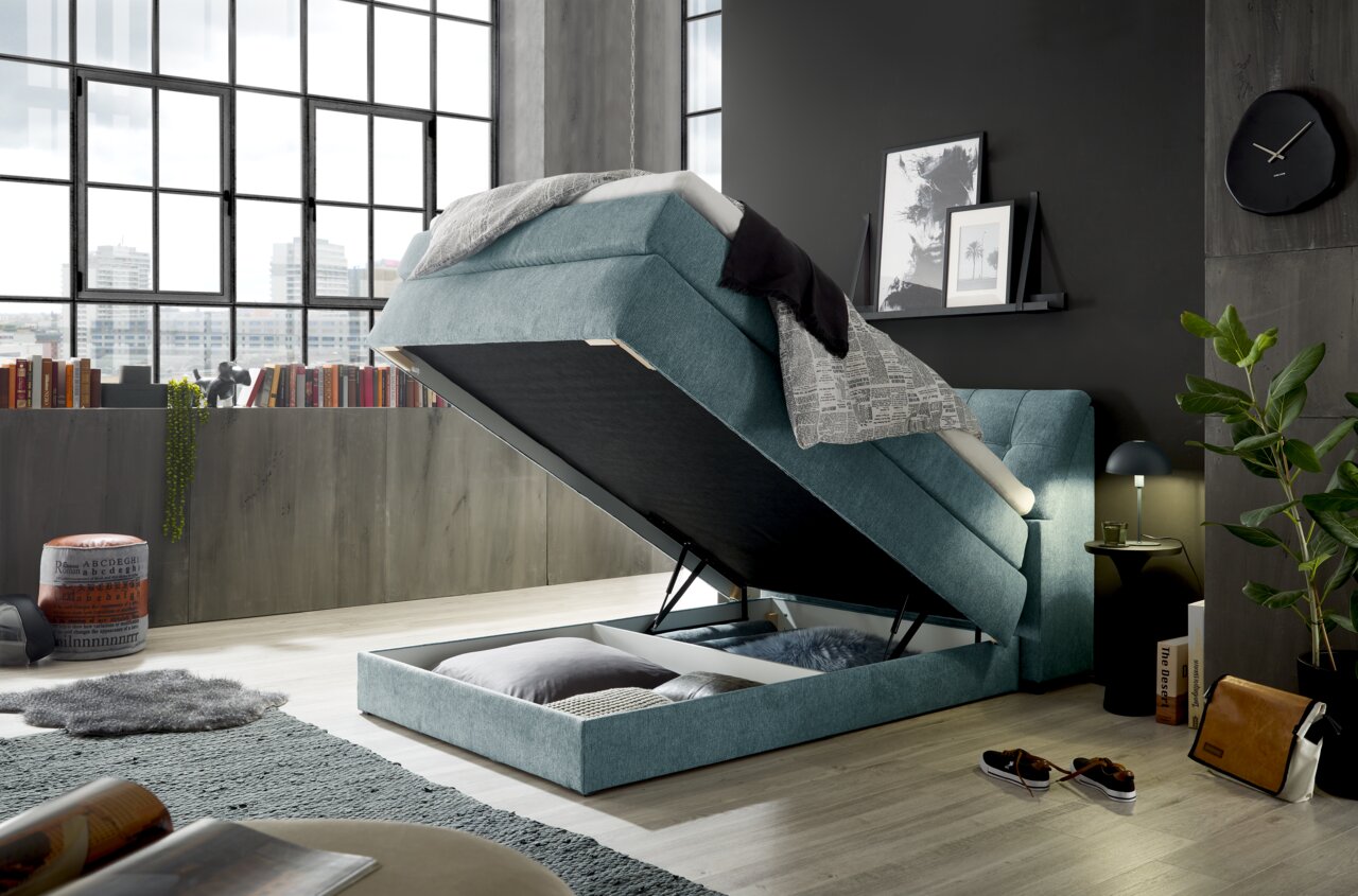 Boxspringbett Set one by Musterring FAIRFIELD in modernem Schlafzimmer, seitliche Perspektive, mit hochgeklappter Matratze und sichtbarem Stauraum darunter.