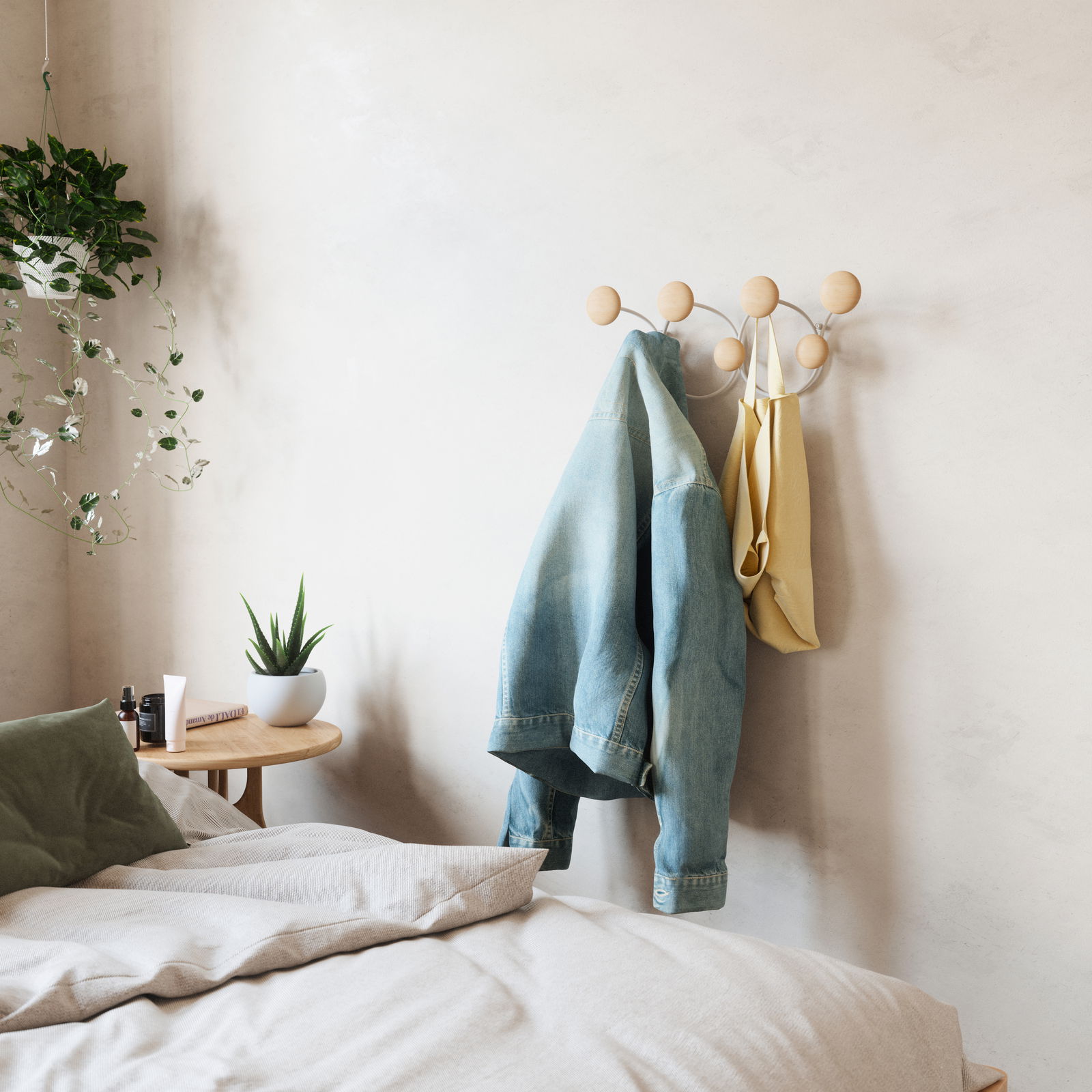 Wandgarderobe mit runden Holzhaken, an der eine Jeansjacke und eine gelbe Tasche hängen, in einem gemütlichen Schlafzimmer mit Pflanzen und einem Nachttisch. Perspektive von der Seite.