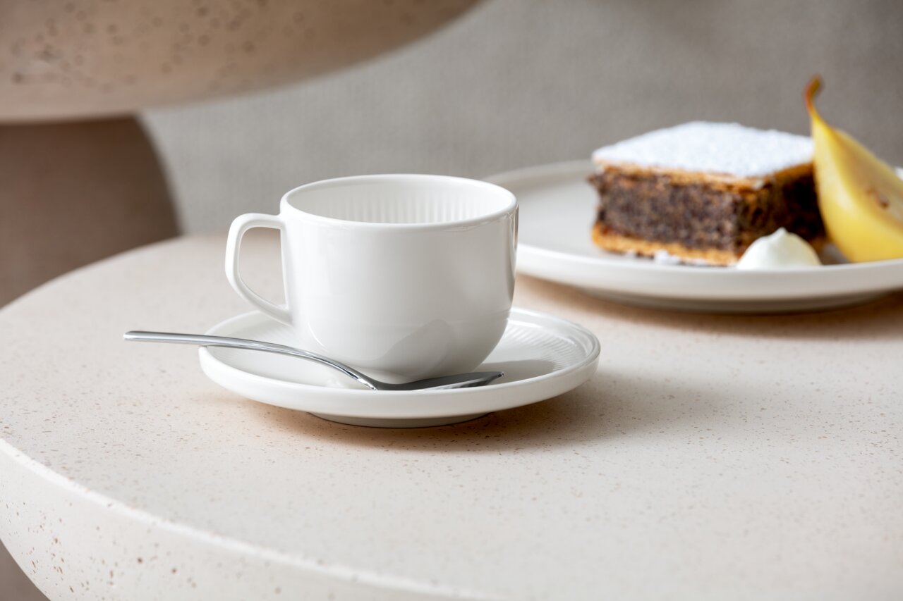 Weiße Kaffeeobertasse Afina 150ml auf Untertasse mit Löffel, seitliche Perspektive, im Hintergrund ein Teller mit Kuchen und Banane.