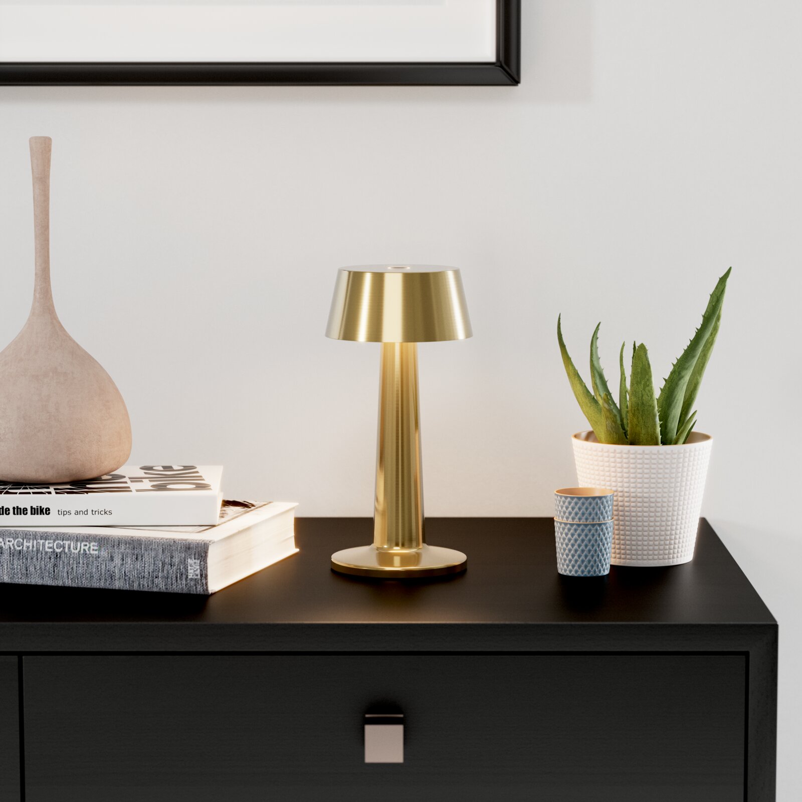 Goudkleurige, moderne tafellamp van metaal op een zwart dressoir, van voren gefotografeerd vanuit een licht verhoogd perspectief, naast boeken, een vaas en een potplant voor een lichte muur