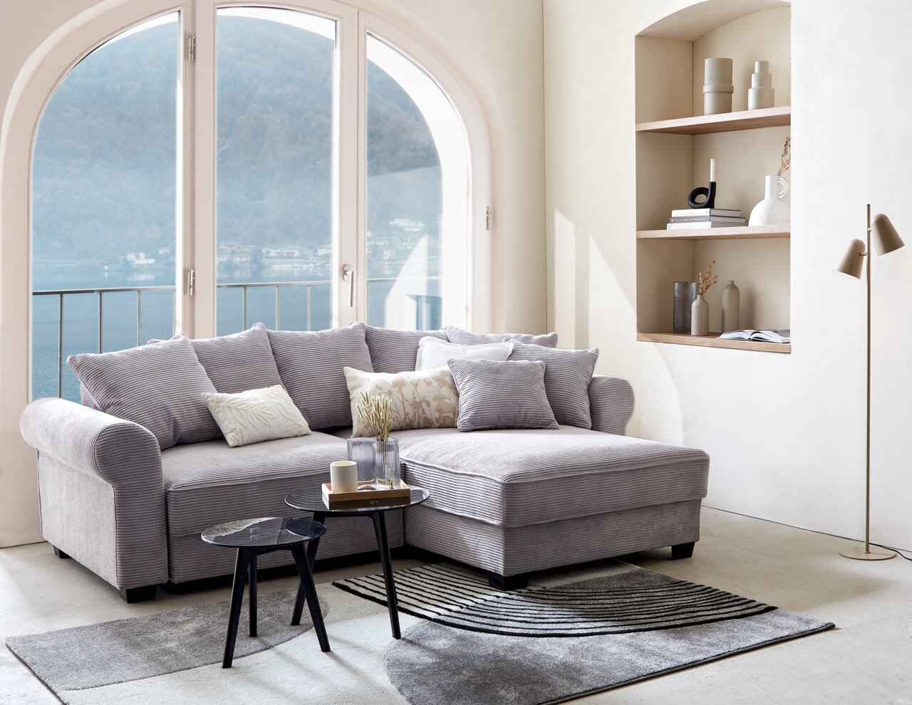 Gemütliche Rundecke MAUREEN in einem hellen Wohnzimmer mit großen Fenstern und Blick auf den See, seitliche Perspektive