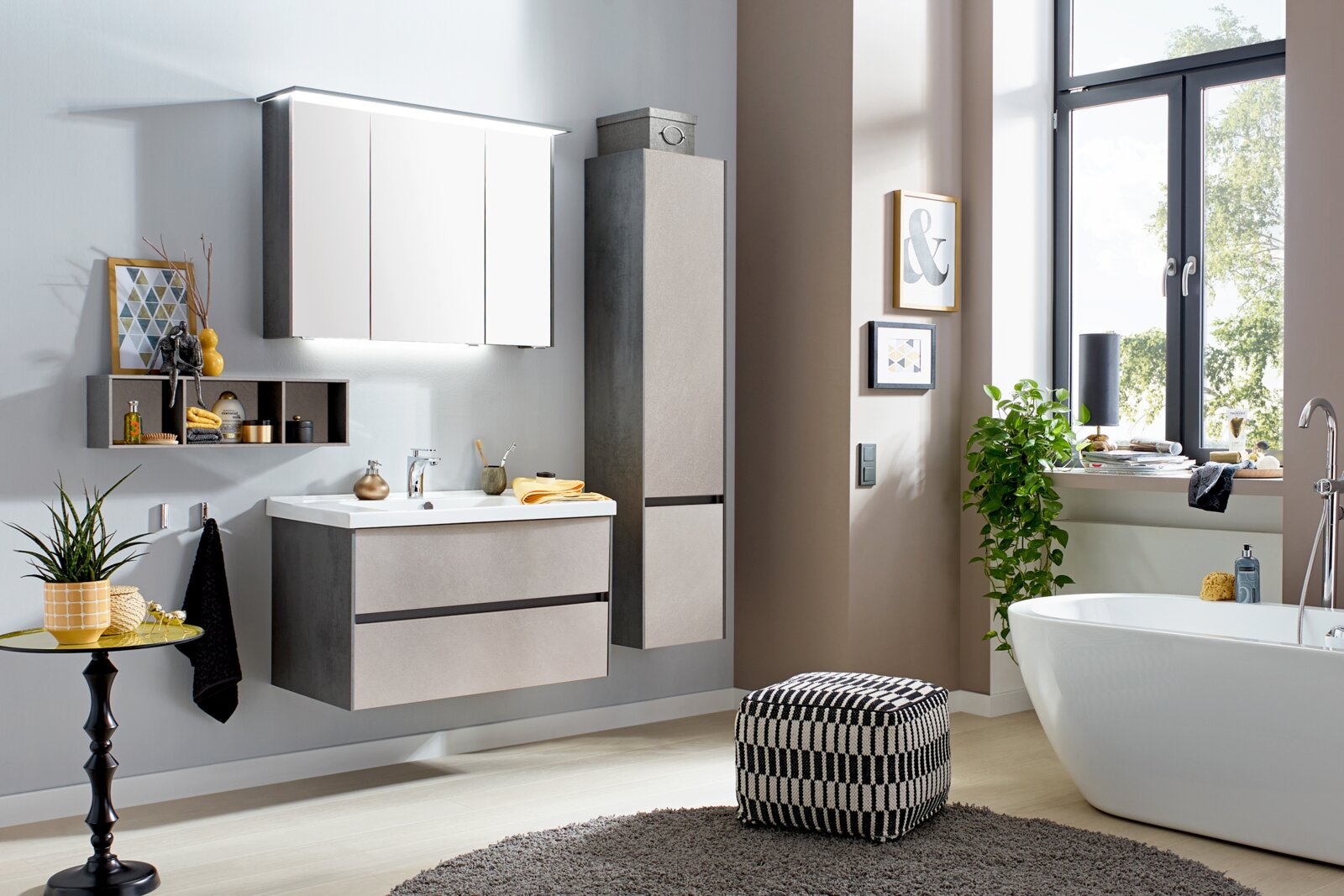 Modernes Badezimmer mit Waschtisch, beleuchtetem Spiegelschrank und Hochschrank in Betonoptik, freistehender Badewanne und großem Fenster, Perspektive: frontale leicht schräge Raumansicht.