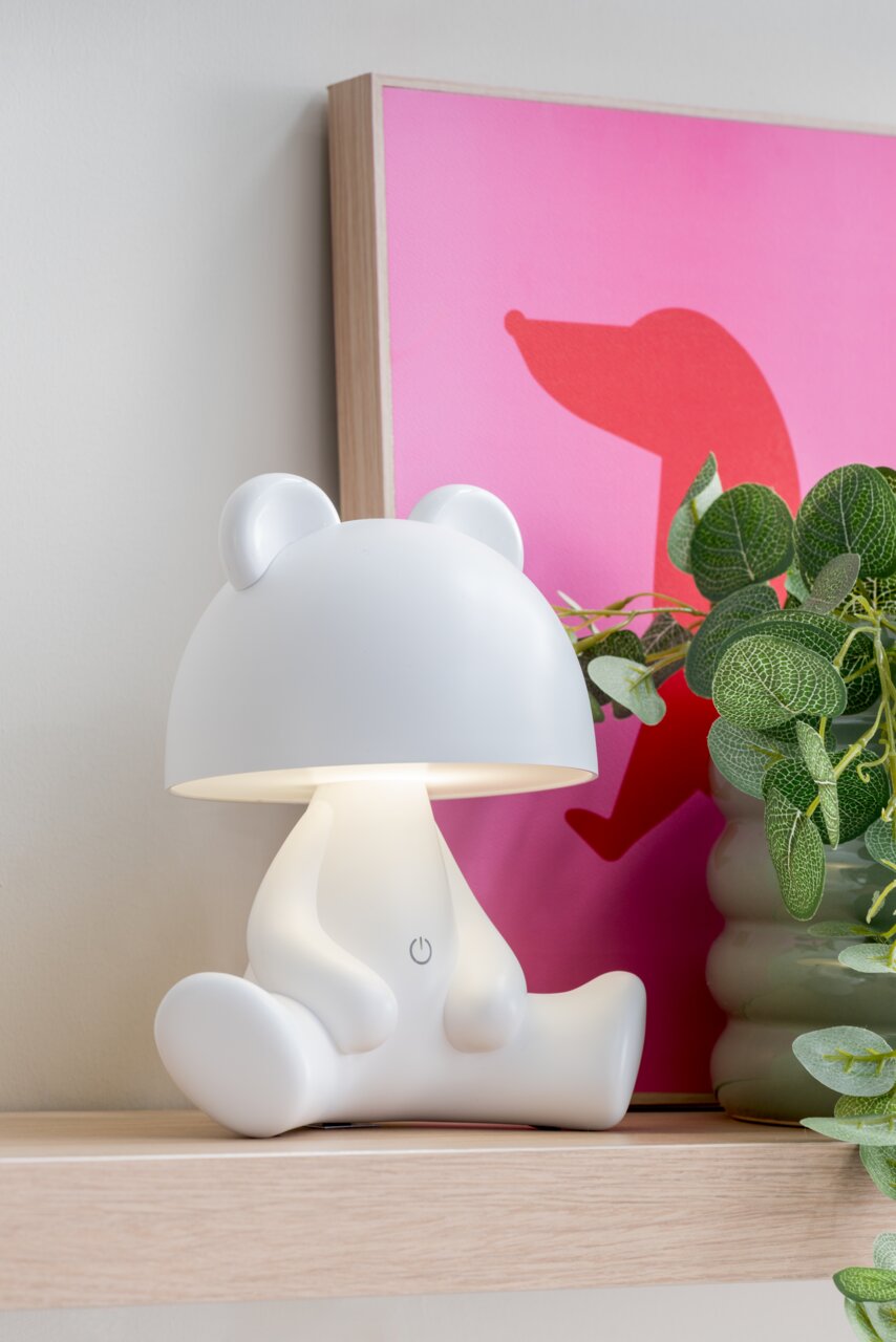 LED-tafellamp in de vorm van een beer op een plank, gezien vanaf de zijkant, naast een plant en een roze schilderij.