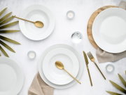 Minimalistisches Tischgedeck mit weißen Porzellantellern und -schalen, goldfarbenem Besteck, Wasserglas und Teelichtern auf hellem Untergrund, von oben fotografiert (Top-Down-Perspektive).