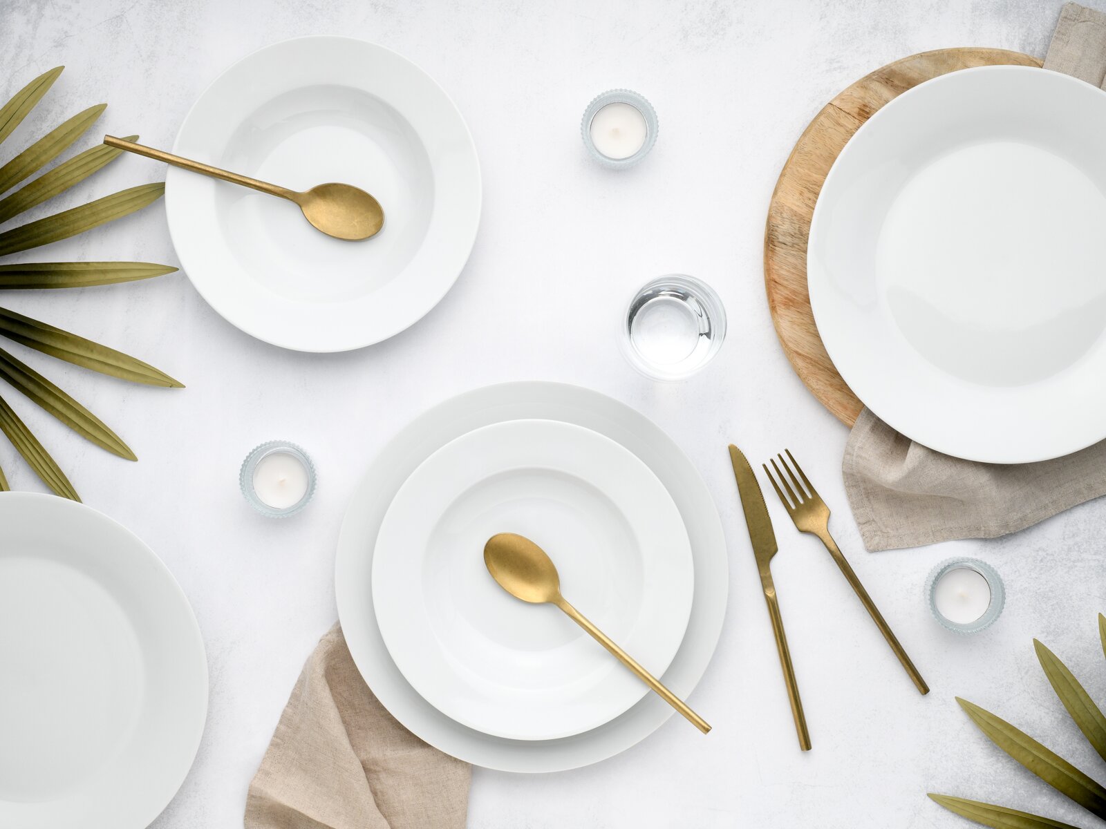 Minimalistisches Tischgedeck mit weißen Porzellantellern und -schalen, goldfarbenem Besteck, Wasserglas und Teelichtern auf hellem Untergrund, von oben fotografiert (Top-Down-Perspektive).