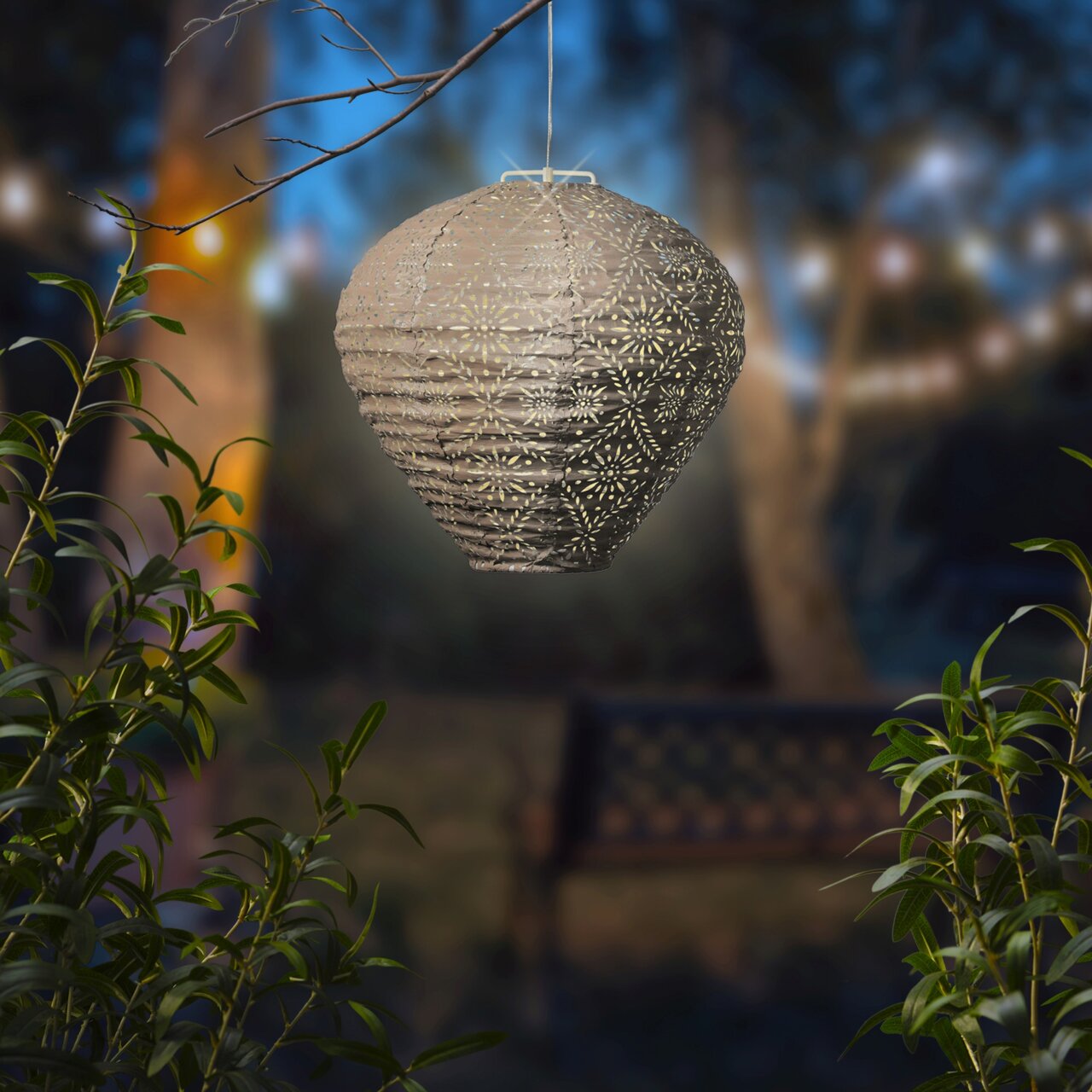 Een hangende zonne-lantaarn in een tuin bij schemering, omringd door planten, met een decoratief patroon en warm licht.