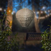 Een hangende zonne-lantaarn in een tuin bij schemering, omringd door planten, met een decoratief patroon en warm licht.