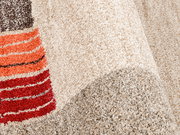 Nahaufnahme eines Teppichs mit beigem Hintergrund und bunten Streifen in Rot- und Brauntönen, perspektivisch von der Seite aufgenommen.