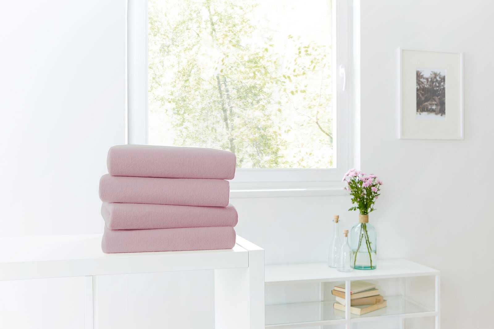 Ein Stapel von vier rosa Spannbetttüchern auf einem weißen Tisch, im Hintergrund ein Fenster mit Blick ins Grüne. Perspektive von vorne.