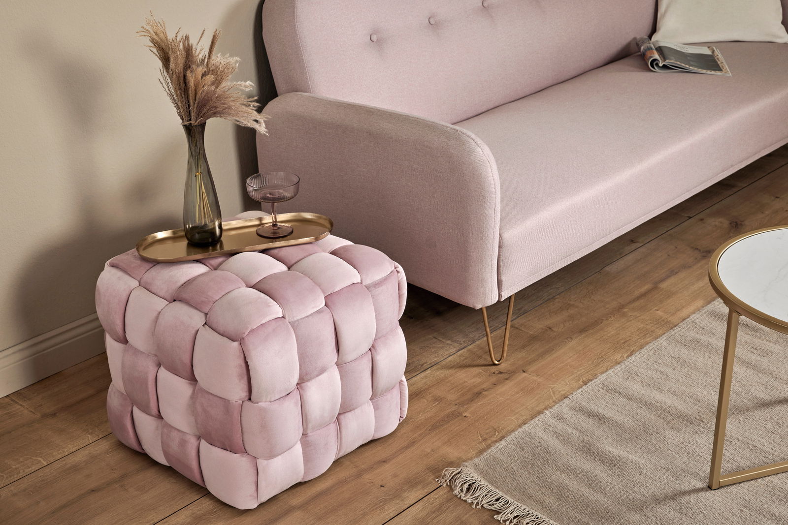 Rosa gepolsterter Hocker in Würfelform mit geflochtenem Design, von schräg oben fotografiert, neben einem Sofa und einem Tisch platziert.