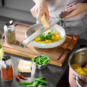 Eine Person reibt Käse mit einer Fein- und Grobreibe über ein Gericht in einer Schüssel. Die Perspektive zeigt die Reibe von oben, während sie über der Schüssel gehalten wird.