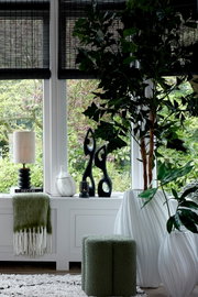 Vooraanzicht van een stijlvolle lichte woonkamer met groene poef, witte geribbelde vazen, zwarte sculpturen, tafellamp, plaid en weelderige kamerplanten voor grote ramen.