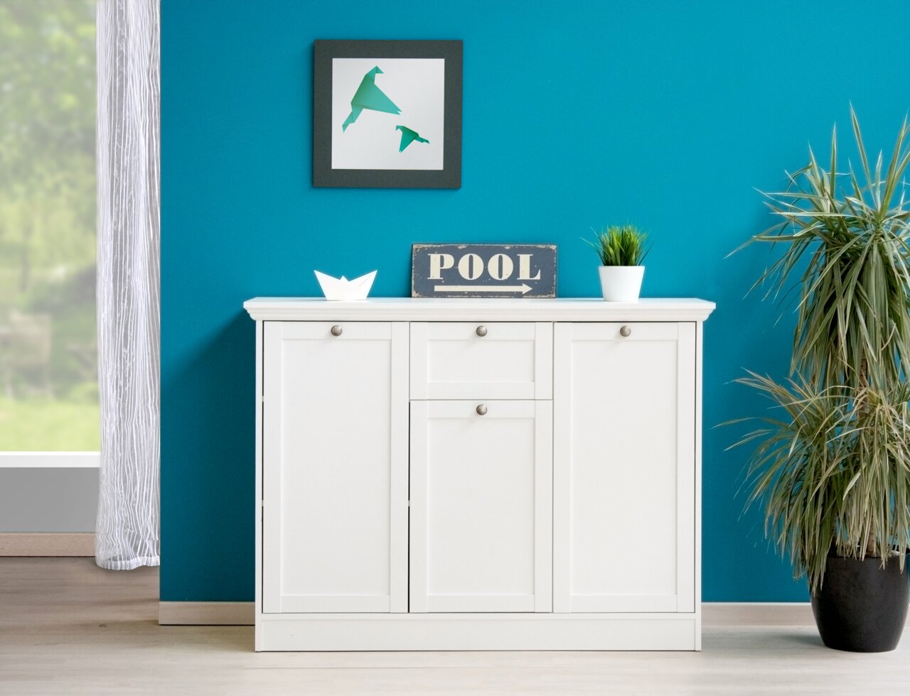 Weiße Kommode mit drei Türen und Schubladen, frontal fotografiert, dekoriert mit Pflanzen und einem Schild auf einer türkisfarbenen Wand.
