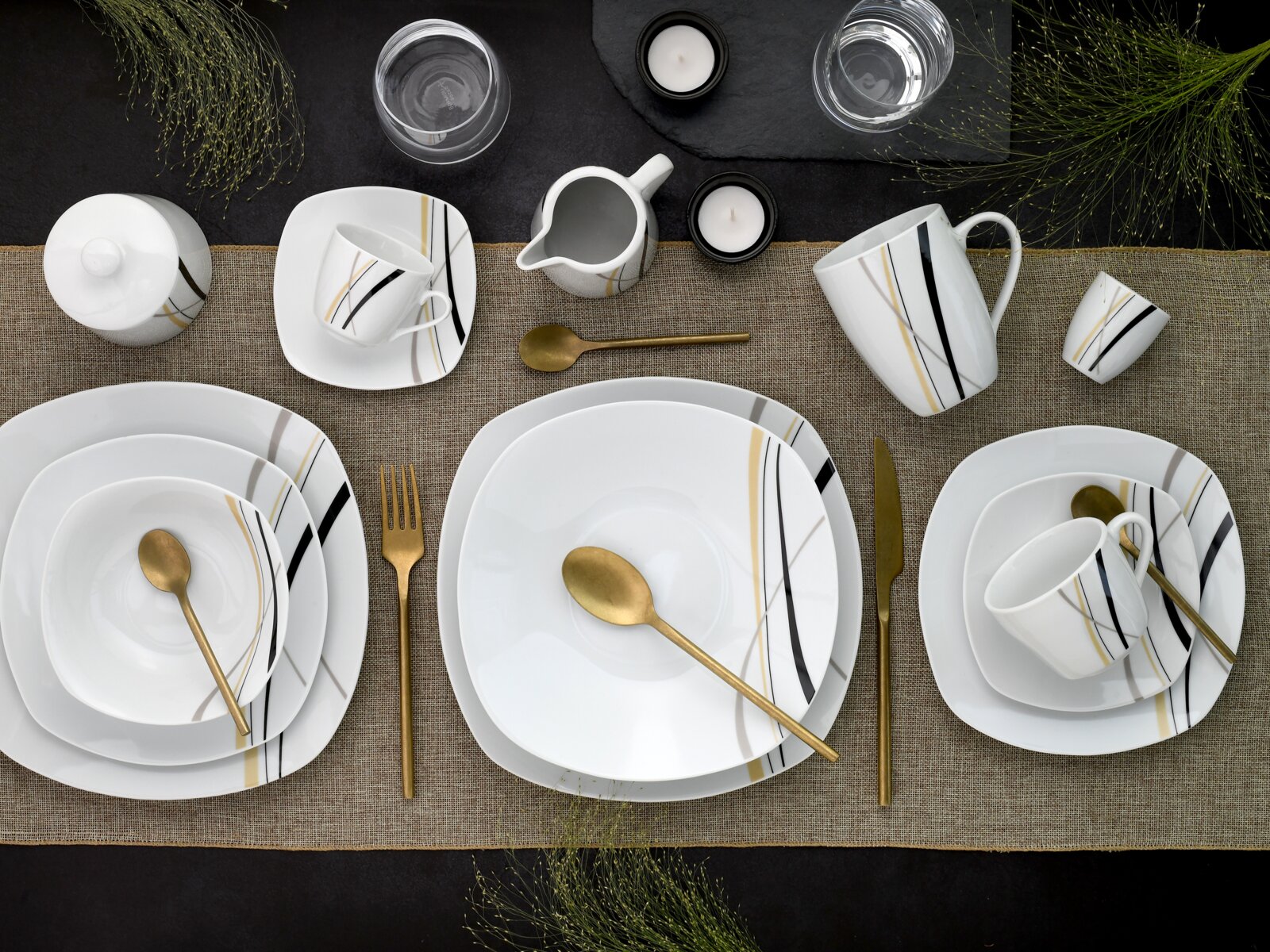 Draufsicht auf ein modern gedecktes Tischarrangement mit weißem Porzellan-Geschirrset (Teller, Schalen, Tassen, Kännchen) mit schwarzen, grauen und goldenen Linien sowie goldfarbenem Besteck auf Leinenläufer, ergänzt durch Gläser und Teelichter.