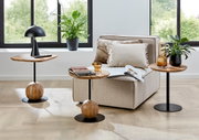 Modernes Wohnzimmer-Setting mit beigem Lounge-Sessel und drei runden Beistelltischen aus Holz und schwarzem Metall, seitliche/frontale Perspektive auf den Sessel und die Tische vor großen Fenstern mit Pflanzen.