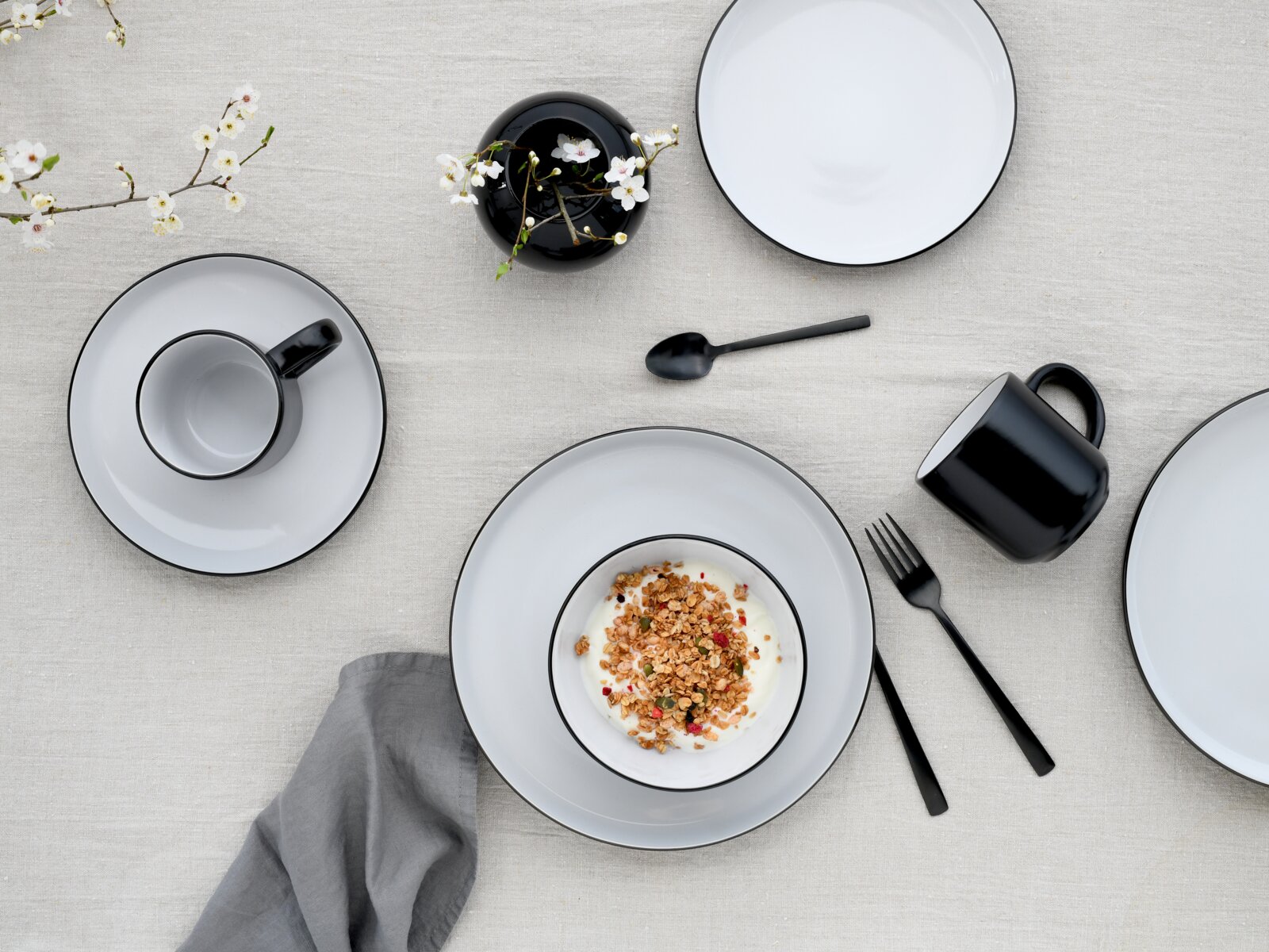 Draufsicht auf ein modern gedecktes Tisch-Arrangement mit weißem Geschirr mit schwarzem Rand, schwarzer Tasse und schwarzem Besteck; in einer Schale Joghurt mit Granola auf hellem Leinentuch, dazu eine schwarze Vase mit Blütenzweigen.