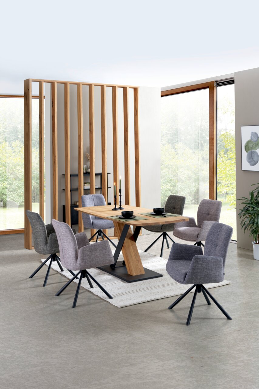 Moderner Esstisch mit Holzplatte und schwarzem Metallgestell, umgeben von sechs grauen Stühlen in einem hellen Raum mit großen Fenstern, aus seitlicher Perspektive.