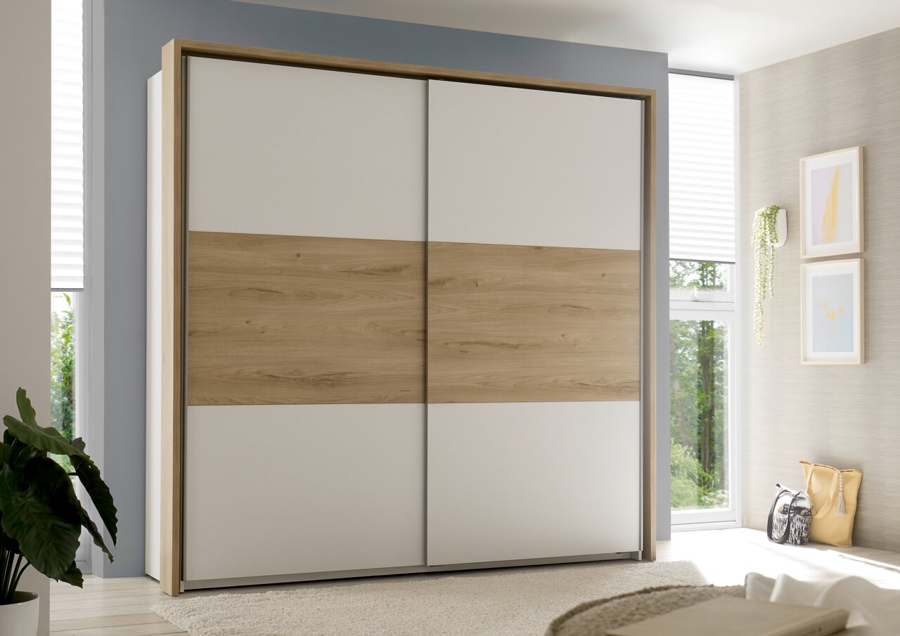 Moderner Kleiderschrank mit Schiebetüren in Holzoptik und weißem Design, frontal fotografiert in einem hellen Schlafzimmer.