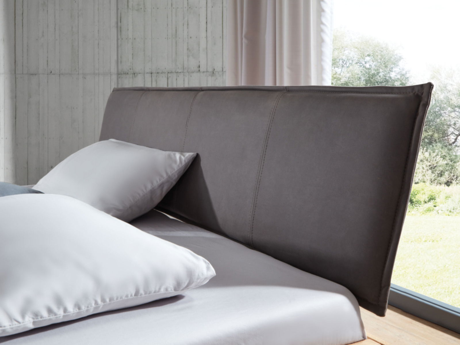 Nahaufnahme eines Balkenbetts mit grauem Kopfteil und weißen Kissen, seitliche Perspektive.