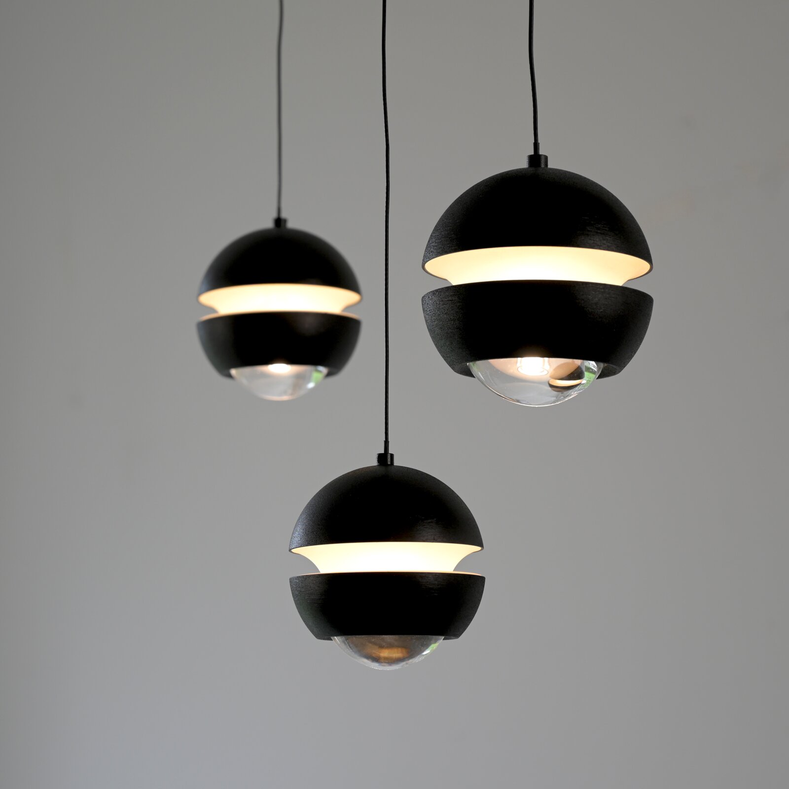 Drei moderne schwarze Kugel-Pendelleuchten mit warmweißem Lichtband und transparentem Glasdiffusor, frontal in leichter Untersicht vor grauem Hintergrund