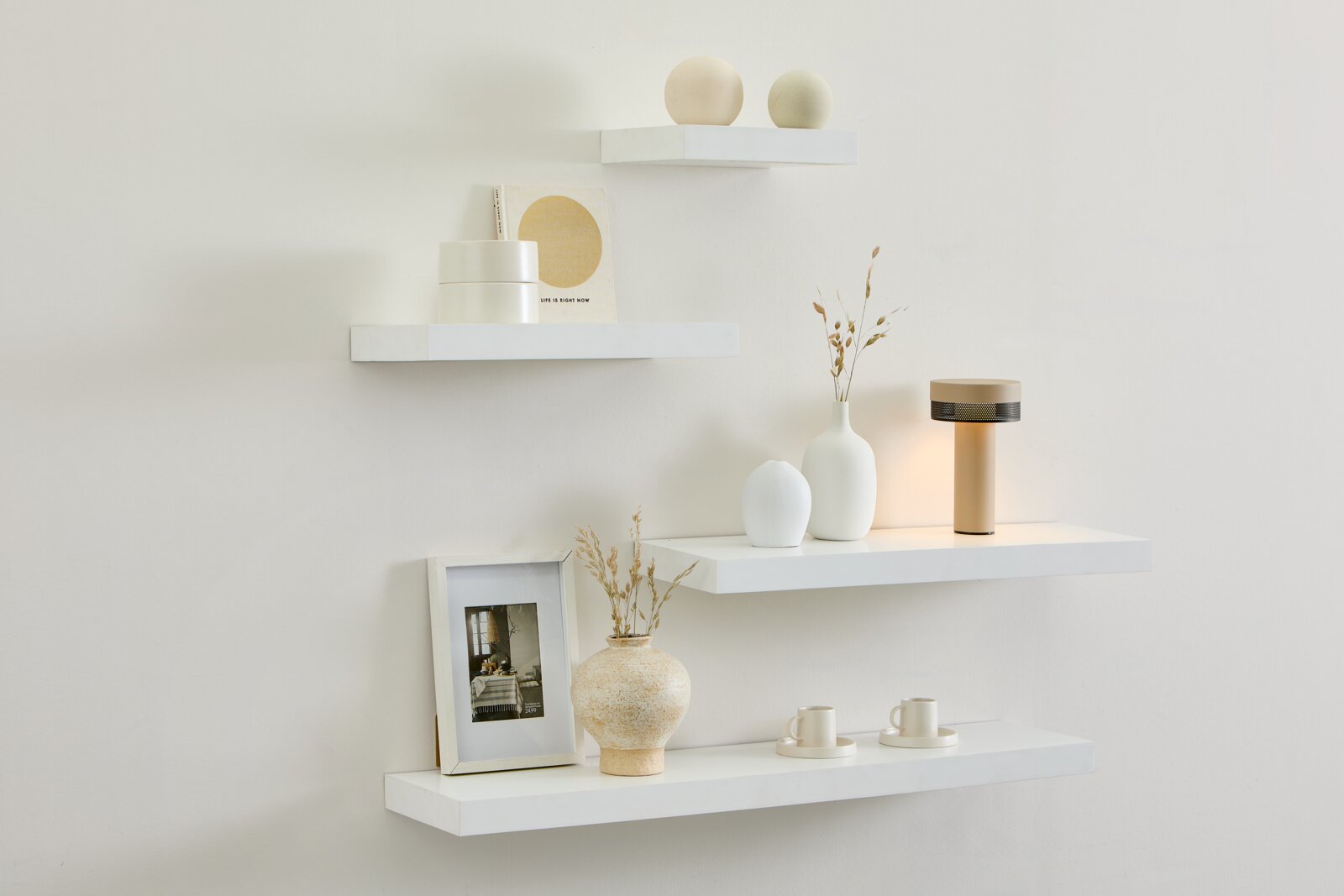 Frontansicht einer stilvoll arrangierten weißen Wand mit vier schwebenden Regalen, minimalistischer Dekoration, Vasen und einer leuchtenden Tischlampe in warmen Naturtönen