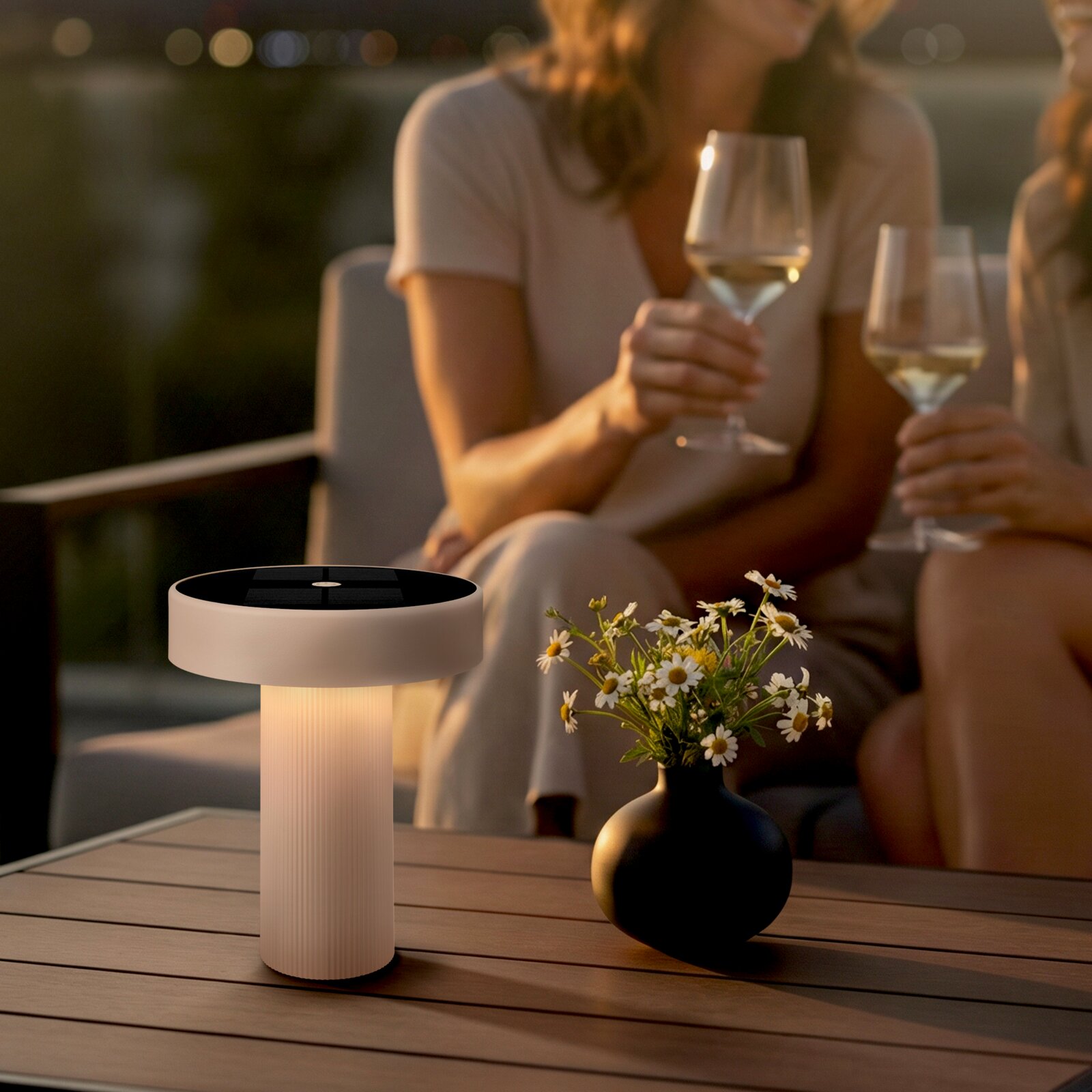Moderne kabellose LED-Tischleuchte mit schwarzer Oberseite und warmweiß leuchtendem, geriffeltem Fuß auf einem Holztisch neben einer Vase mit Gänseblümchen; Perspektive: frontale Nahaufnahme auf Tischhöhe, leicht von links.
