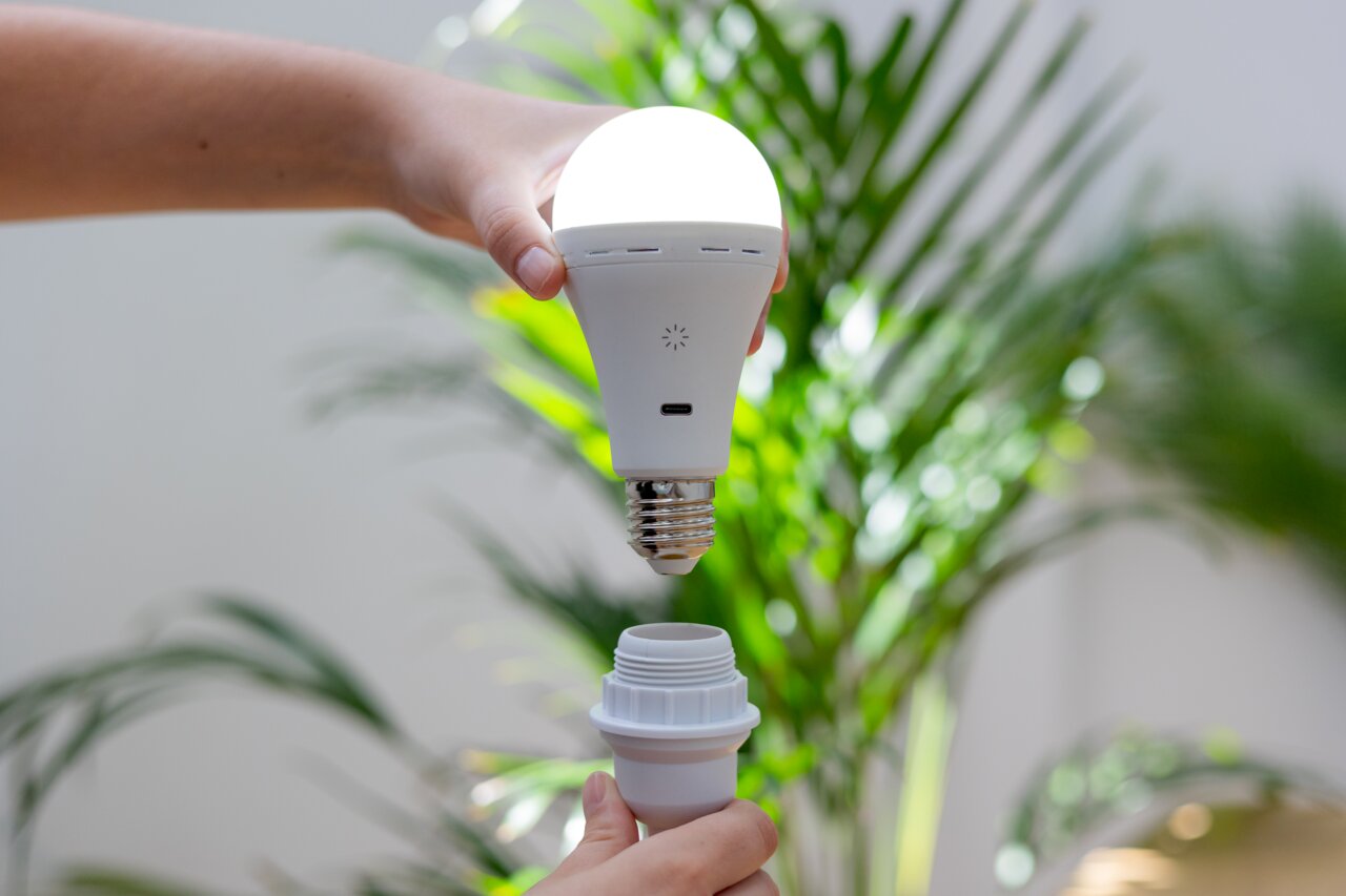 Zijaanzicht: een hand houdt een oplichtende oplaadbare lamp boven een fitting, met op de achtergrond groene planten.