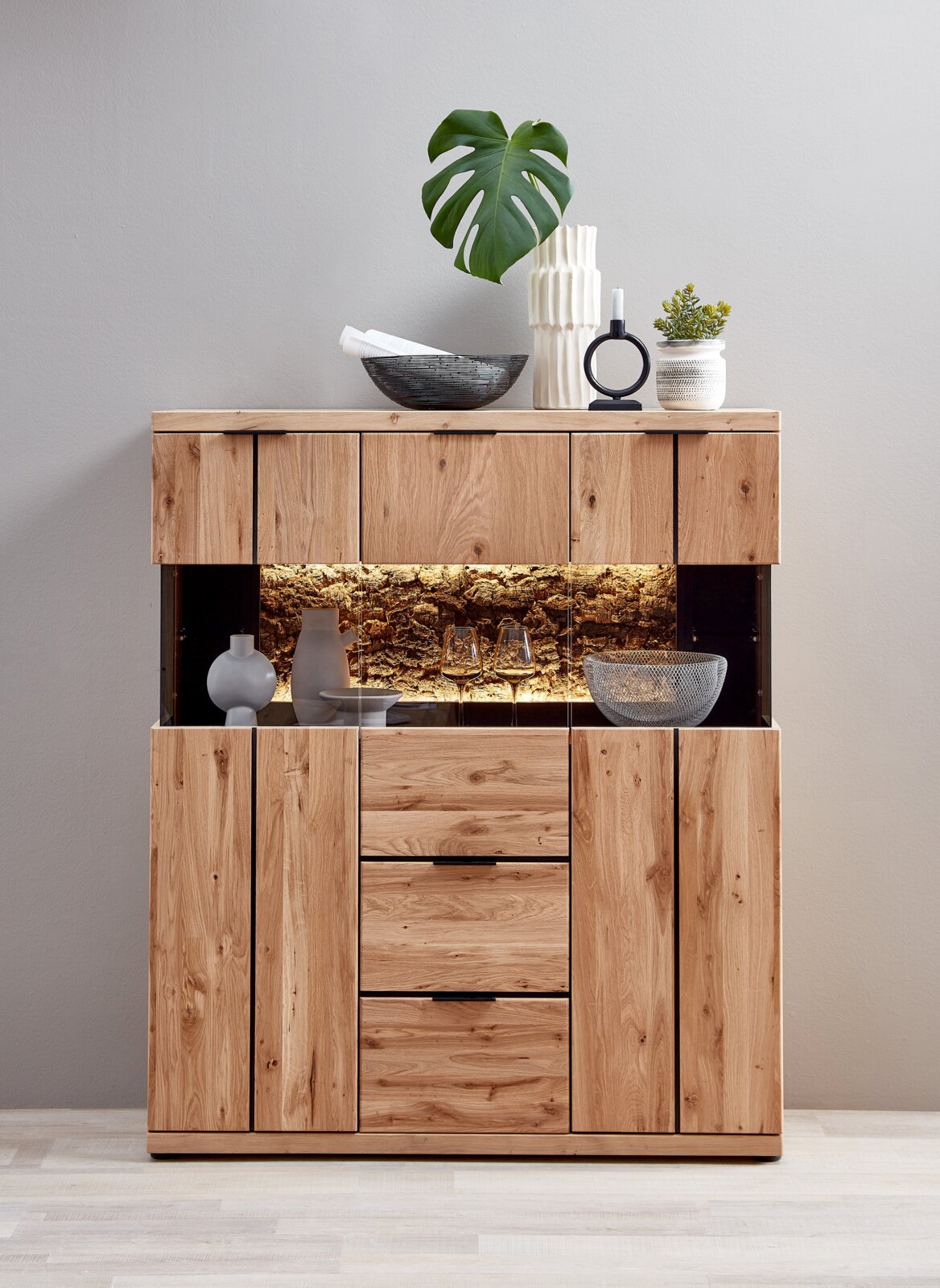 Cantus Highboard DONNA Frontansicht eines modernen Highboards aus massivem Holz mit Glasvitrine und warm beleuchtetem Naturkork-Rückwandbereich, dekoriert mit Vasen, Schalen, Kerzenhalter und Pflanze auf hellem Hintergrund