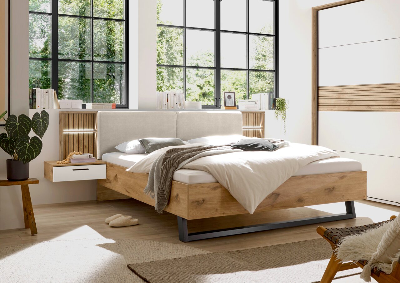Z2 Bettanlage DARIA/LEO Modernes Schlafzimmer mit einem stilvollen Bett aus Holz und gepolstertem Kopfteil, seitlicher Perspektive. Das Bett ist von natürlichem Licht durch große Fenster umgeben, mit einem Nachttisch und dekorativen Elementen.