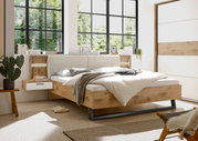 Modernes Schlafzimmer mit einem stilvollen Bett aus Holz und gepolstertem Kopfteil, seitlicher Perspektive. Das Bett ist von natürlichem Licht durch große Fenster umgeben, mit einem Nachttisch und dekorativen Elementen.